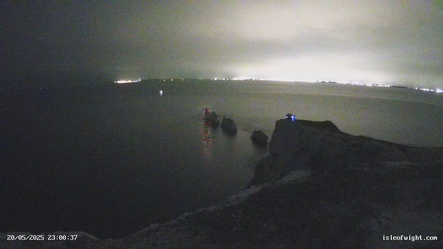 A nighttime scene of a rocky coastline with a calm body of water in the foreground. There are large rocks on the shoreline, with some structures or equipment visible on the rocks, possibly lights or sensors. In the background, the horizon shows distant city or town lights against a cloudy sky illuminated by a bright glow, likely from the city or weather conditions. The timestamp in the bottom left corner indicates the date and time as May 20, 2025, at 23:00:37. The URL "isleofwight.com" is visible in the bottom right corner.