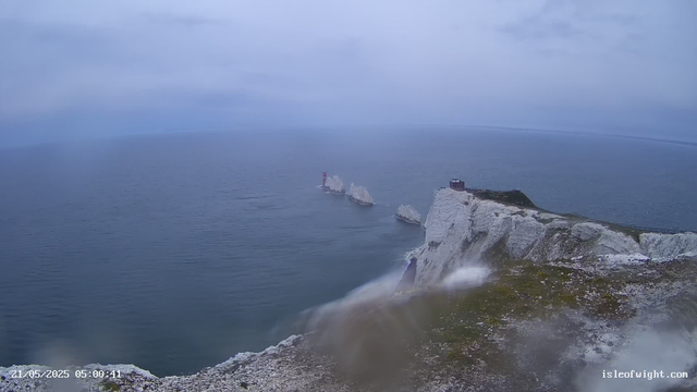 A view of white chalk cliffs with a signal tower and a small building on top, overlooking the ocean. Waves are crashing against the base of the cliffs, and the sky is overcast with gray clouds. A red and white lighthouse is visible on one of the distant rocks in the water. The scene appears to be during early morning or late evening, with a timestamp of 21/05/2025 05:00:41. The website "isleofwight.com" is displayed in the bottom right corner.