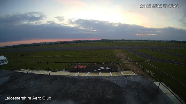 A wide view from a webcam showing an early morning sky with clouds and the horizon in the distance. In the foreground, there is a helipad and some fencing, with open grassy fields and a runway extending into the background. The date and time stamp in the upper right corner indicates it is 5:00 AM on May 21, 2025. The bottom left corner displays the text "Leicestershire Aero Club."