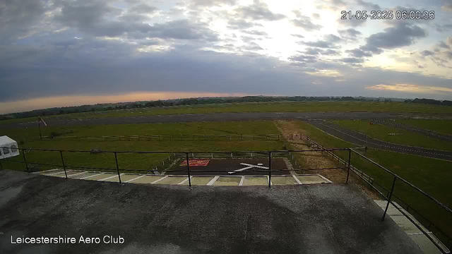 A panoramic view of an open landscape taken from a high vantage point at dawn or dusk. The sky is partly cloudy with patches of sunlight breaking through the clouds. In the foreground, there is a flat rooftop with a railing, a small helipad painted with an "H" and a red border, and a parking area with marked spaces. The midground features a grassy field enclosed by a fence, with a small structure and a windsock on a pole. In the distance, there are open fields extending toward a horizon with a few scattered trees. The image is labeled "Leicestershire Aero Club" and has a timestamp of "21-05-2025 06:00:36" in the upper right corner.