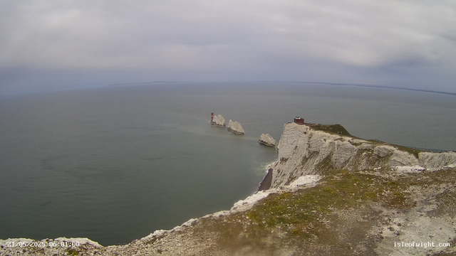 A view of white chalk cliffs over the sea on a cloudy day, with several large rocks jutting out of the water near the base of the cliffs. A small building and a lighthouse are visible on top of the cliffs. The horizon shows a distant shoreline under an overcast sky.