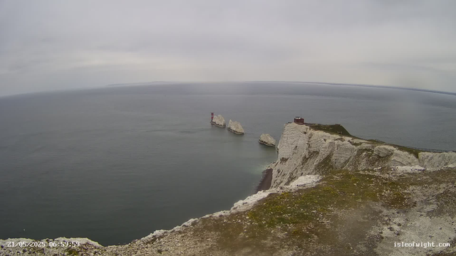A view of white chalk cliffs extending into a calm, gray sea under an overcast sky. Three small rocky islands with a red and white lighthouse are visible near the cliffs. The land at the top of the cliffs is grassy with a small structure and a fence along the edge.