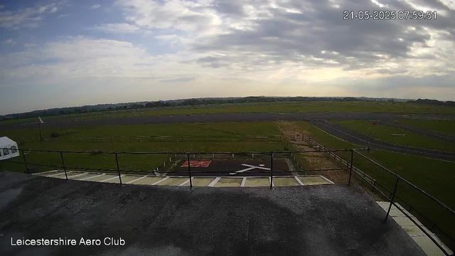 An outdoor view of an airfield or race track under a partly cloudy sky, with a grassy landscape extending into the horizon. In the foreground, there is a platform or balcony with a black railing and a helipad marked with an "H". The timestamp "21-05-2025 07:59:51" is displayed in the top right corner, and the text "Leicestershire Aero Club" appears at the bottom left.