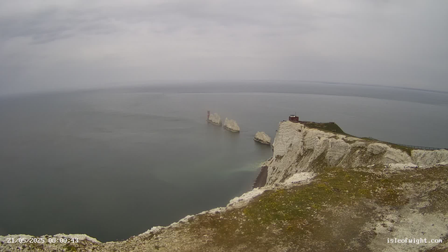 A panoramic view of white chalk cliffs rising sharply from the sea under a cloudy sky. Four prominent rock formations and a lighthouse are visible near the edge of the cliffs. The ocean appears calm, and the landscape has patches of grass and moss on the cliff tops. The scene is overcast, giving a muted tone to the entire view. The timestamp indicates the photo was taken on May 21, 2025, at 08:00:43.
