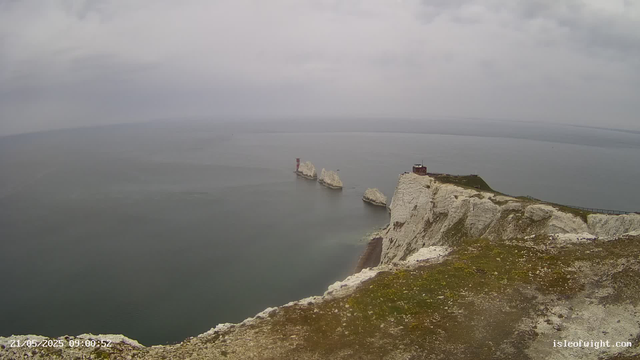 A view of white chalk cliffs extending into the sea under a cloudy sky. Several large chalk stacks are visible offshore, and there is a small lighthouse or navigational marker on one of the stacks. The cliffs are rugged and steep, with some grassy patches on top. The water appears calm, and the landscape has a misty, overcast atmosphere.