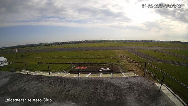 An outdoor view from a webcam at Leicestershire Aero Club shows a grassy airfield with runways stretching into the distance. The sky is partly cloudy with sunlight shining through, casting a bright scene. In the foreground, there is a small helipad marked with a white "H" on black, surrounded by a railing, and adjacent parking spaces. The timestamp in the top right corner indicates the date as May 21, 2025, and the time as 09:00:49.