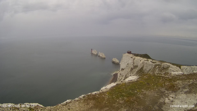 A view of white chalk cliffs extending into the sea under a cloudy sky. Several large sea stacks rise from the water near the cliffs, with a small red lighthouse or navigation tower atop one of the stacks. The water appears calm, and a narrow path runs along the edge of the cliffs. The landscape is misty and overcast.