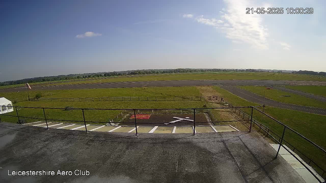A wide-angle view from a webcam overlooking a flat, grassy outdoor area with a small paved section and a metal railing in the foreground. In the distance, there are runways or paved pathways, with a partly cloudy sky overhead. The time and date displayed are 21-05-2025 at 10:00:29. The location indicated is Leicestershire Aero Club.