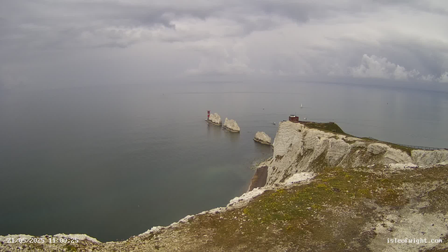 A view of the white chalk cliffs of the Isle of Wight under a cloudy sky, with the Needles lighthouse and rock formations visible in the distance, overlooking the calm sea.