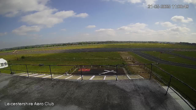 A view from a webcam showing an outdoor area of Leicestershire Aero Club. In the foreground, there is a paved deck with a metal railing and several empty parking spaces. Beyond the deck, there is a grassy field with a small red and black helipad marked with a white helicopter symbol. The background features open green fields and a blue sky with scattered white clouds, and the scene appears to be taken on a sunny day. The timestamp on the top right corner reads "21-05-2025 11:00:48."