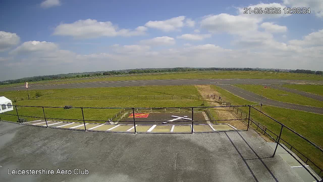 A wide view of an airfield taken from a webcam. The image shows a grassy area with a runway extending into the distance. The sky is partly cloudy with scattered white clouds and a bright blue background. There is a small, fenced-off area with a helipad marked with a red and yellow symbol. In the foreground, there is a parking area and a white vehicle. The timestamp in the top right corner indicates the date and time as May 21, 2025, at 12:00:04. The bottom left corner displays the text "Leicestershire Aero Club."