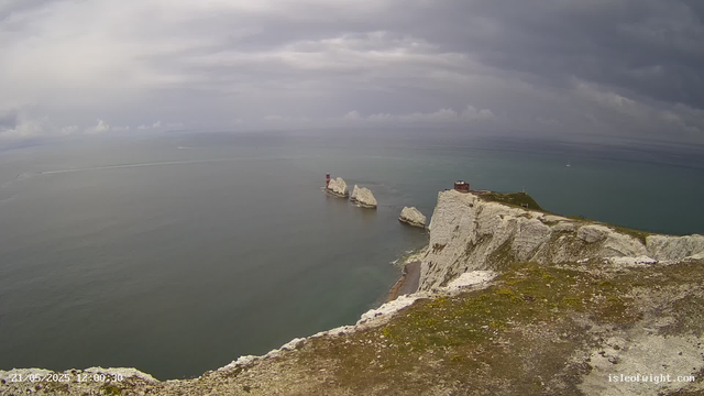 A view from a high vantage point over white chalk cliffs extending into the ocean. The cliffs are topped with green grass, and there is a small building or structure near the edge. Several large rock formations are visible jutting out of the water near the base of the cliffs. The sky is cloudy with patches of darker clouds, and the sea appears calm. A small boat can be seen in the distance, leaving a white trail behind it. The timestamp on the image reads "21/05/2025 12:00:30," and the watermark "isleofnight.com" is visible in the lower right corner.