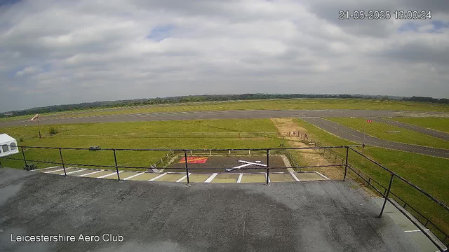 A view from a webcam at LeicesterShire Aero Club showing an airport runway and surrounding grassy area. The sky is partly cloudy with patches of blue, and the timestamp reads 21-05-2025 13:00:24. There are parking spaces visible in the foreground, some marked for designated use, and a fenced-off area near the parking lot. The runway extends into the distance with some markings, and a small wind turbine can be seen on the grassy field to the left. The horizon shows a line of trees or low hills under a mostly cloudy sky.