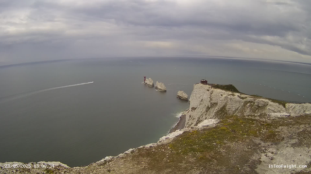 A view from a cliff overlooking the ocean under a cloudy sky. There are large white chalk formations and a small building on the edge of the cliff, with a lighthouse standing on one of the rocks in the water below. A boat is visible in the distance leaving a wake behind it. The coast extends along the horizon, and the scene has a cool, overcast atmosphere.