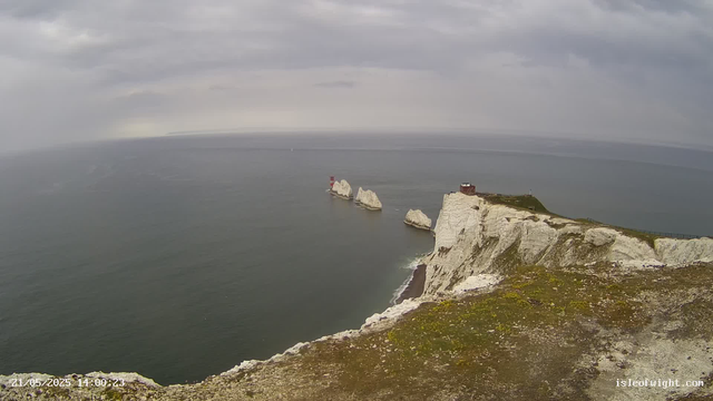 A view of white chalk cliffs with patches of green moss, extending into the ocean under a cloudy sky. There are several large rock formations in the water near the base of the cliffs, and a small building or lookout at the top of the highest cliff. The sea appears calm with gentle waves, and the horizon stretches into the distance beneath a grey, overcast sky.