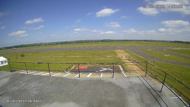 A clear daytime view of an airfield with a runway extending into the distance, surrounded by green fields under a blue sky with scattered clouds. In the foreground, there's a balcony with a black railing, and below it, a small designated area with painted markings and a red rectangular object. The image has a timestamp in the upper right corner showing "21-05-2025 14:00:56" and the text "Leicestershire Aero Club" in the lower left corner.