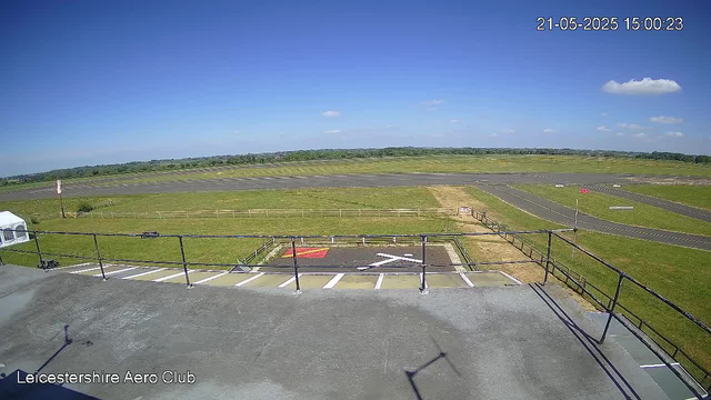 A view of an airfield with a clear blue sky and scattered clouds. In the foreground, there is a parking area with multiple empty parking spaces, a helipad marked with a red and white "H", and a black railing. Beyond the parking area, there is a grassy field leading to a paved runway with markings. A small sign is visible near the fence, and a white tent is partially visible on the left side. The timestamp in the top right corner shows the date and time as 21-05-2025 15:00:23. The bottom left corner has text reading "Leicestershire Aero Club."