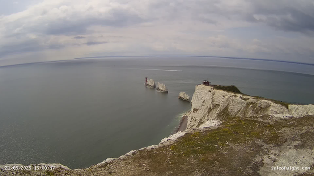 A view from a webcam showing white chalk cliffs overlooking the sea. In the distance, three large rock formations are visible in the water, with a lighthouse standing on each of two of them. A pathway runs along the top of the cliffs, and there is a small building on the cliff edge. The sky is partly cloudy with patches of blue. A boat is moving across the water, leaving a trail behind. The timestamp in the lower left corner reads "21/05/2025 15:00:47," and the bottom right corner has the website "isleofwight.com."