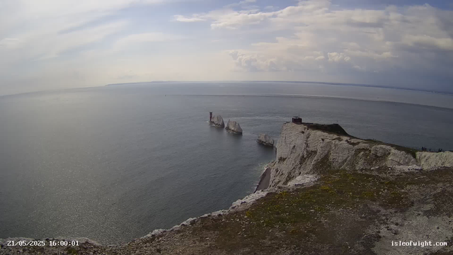 A panoramic view of white chalk cliffs with a grassy top, overlooking a calm sea under a partly cloudy sky. Three prominent rock stacks rise out of the water near the cliff base. The coastline extends into the distance on the horizon.