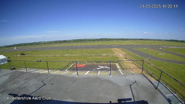 The image shows a wide view of an airfield taken from a webcam, with a clear blue sky overhead. In the foreground, there is a parking area with marked parking spaces and a small helipad. A white tent is visible on the left side. The runway stretches across the middle distance with green grassy areas surrounding it. In the background, the horizon features a line of distant trees and open fields. The top right corner displays the date and time: 21-05-2025 16:00:11. The bottom left corner has the text "Leicestershire Aero Club."
