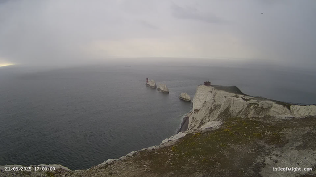 A view of white chalk cliffs rising from the ocean on a cloudy day. There are several large rocks near the base of the cliffs in the water, and a small building or lighthouse is situated on top of the highest point in the cliffs. The sky is overcast, and the sea appears calm. A faint horizon line can be seen in the distance. The image is timestamped "21/05/2025 17:00:00" and labeled with the website "isleofwight.com".