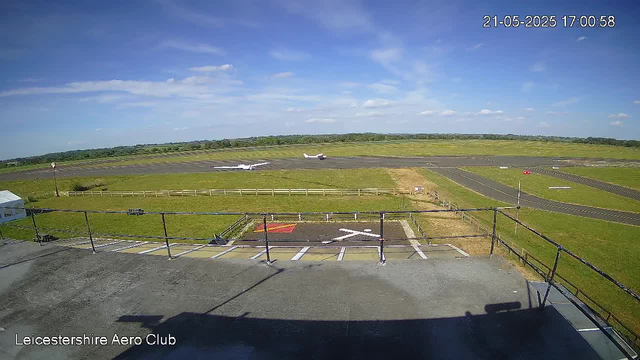 A view of a small airport runway on a sunny day, with two small airplanes taxiing on the tarmac. The sky is mostly clear with a few scattered clouds. In the foreground, there is a helipad marked with an "H" and a parking area. The background features green fields and a distant horizon. The timestamp indicates the date and time as 21st May 2025 at 17:00:58. The bottom left corner has the text "Leicestershire Aero Club."