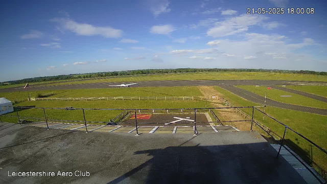 A wide view of an airfield under a blue sky with scattered clouds. In the foreground, there is a concrete rooftop with a safety railing, and a helipad logo painted on the surface. A small airplane is parked on the grass near the runway, with its nose pointing toward the horizon. The runway stretches out into the distance, surrounded by grassy fields and a distant tree line. The date and time are displayed in the top right corner, and the text “Leicestershire Aero Club” is visible in the bottom left corner.