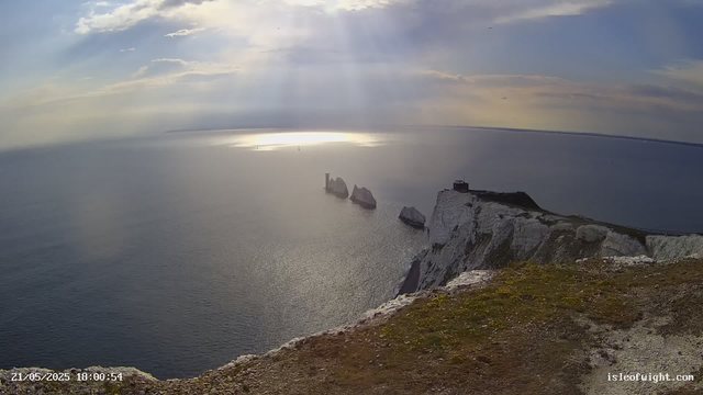 A high-angle view of white chalk cliffs on the coastline extending into the ocean. The cliffs have a rugged, steep drop into the sea below. The sky is partly cloudy with sunlight breaking through, creating a reflection on the water. There are three prominent rock stacks offshore near the cliffs. A small structure is visible on top of the highest part of the cliffs. The foreground shows a grassy and rocky edge of the cliff. The timestamp in the corner reads "21/05/2025 18:00:54," and the bottom right corner has the website "isleofwight.com."