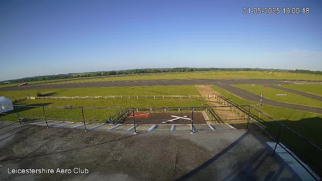 A clear view of a tarmac runway and grassy fields beyond, with a blue sky overhead. The image captures part of an airfield at Leicestershie Aero Club, with white racing lines painted on the ground and a sign indicating a runway. The time and date are shown in the top right corner as "21-05-2025 19:00:18." The scene is bright and sunny with no visible clouds.