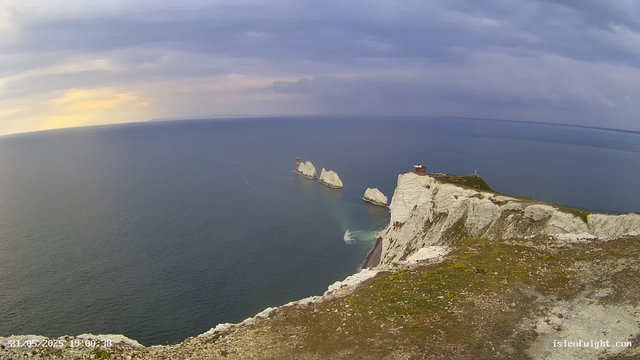 A view of white chalk cliffs rising sharply from the ocean with a grassy top, leading to a small building and a lighthouse. Three large rock formations are visible in the water near the cliffs. The sky is partly cloudy with patches of yellow and purple hues, indicating either sunrise or sunset. The date and time displayed in the corner is "21/05/2023 19:00:38."