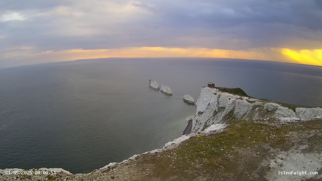 A view from a webcam showing white chalk cliffs overlooking the sea at sunset. The sky is partly cloudy with a mix of dark clouds and a golden glow near the horizon. There are several large chalk rock formations just off the coast, and a small building perched atop the cliff. The foreground contains rocky, grassy terrain. The timestamp on the image reads "21/05/2025 20:00:51," and the website "isleofwight.com" is displayed in the bottom right corner.