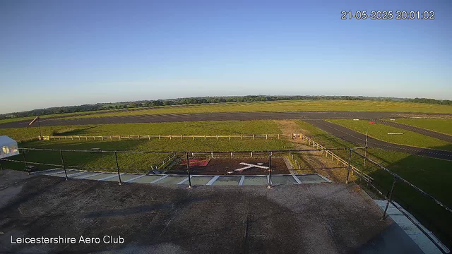 A wide view of an airfield taken from a webcam, showing a runway in the background, open grassy areas, and a small helicopter or drone landing pad in the foreground. The sky is clear and blue, and the scene is brightly lit by sunlight. The timestamp on the image indicates it was captured on May 21, 2025, at 20:01:02, and there is a label in the bottom left corner that reads "Leicestershire Aero Club."