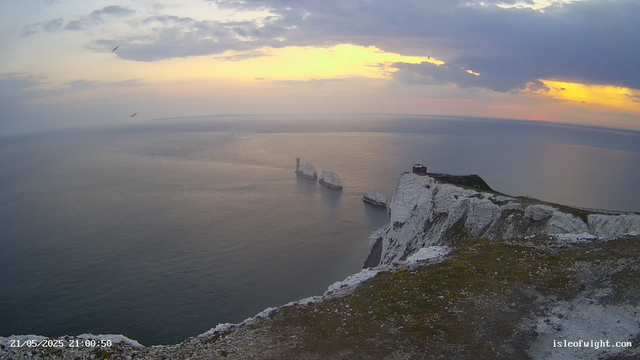 A view from a webcam showing the white chalk cliffs of the Isle of White at sunset. The sky is partly cloudy with hints of orange and yellow near the horizon. The sea is calm, and several ships are visible near the base of the cliffs. The timestamp in the lower left corner indicates the date and time as May 21, 2025, at 9:00 PM. The website "isleofwight.com" is in the lower right corner.