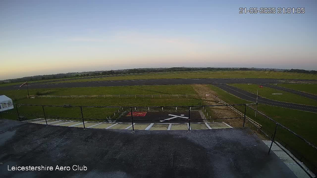 A wide view of an airfield at sunset, with a small helicopter landing pad in the foreground marked with a white "H" and red circle, surrounded by safety fences. The runway and grassy areas extend into the distance under a clear sky, with the timestamp "21-05-2025 21:01:05" in the top right corner. The bottom left corner displays the text "Leicestershire Aero Club."