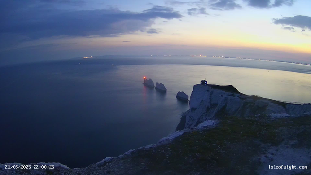 A view of the white chalk cliffs at sunset, with a lighthouse on a prominent cliff in the foreground. In the distance, the sea is calm with a few boats and a faint horizon line. The sky has scattered clouds, and the overall scene is serene and tranquil. The timestamp in the bottom left corner shows "21/05/2025 22:00:25," and the website "isleofwight.com" is displayed in the bottom right corner.