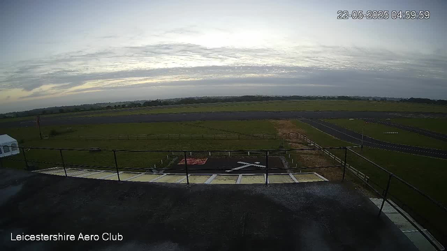 An outdoor view from a webcam at Leicesterhire Aero Club shows a cloudy sky with the sun either rising or setting on the horizon. In the foreground, there is a rooftop with a safety railing, and a paved area with markings and a small helipad. The scene extends over a grassy field with pathways and some distant trees or structures on the horizon. The timestamp in the top right corner indicates the date and time as May 22, 2025, at 4:59 AM or PM.