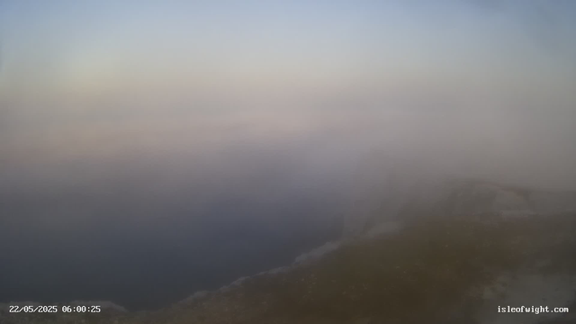 A foggy, overcast coastal scene at dawn or early morning. The image shows the shoreline with waves crashing against the rocks, and a hazy sky with very little visibility. The timestamp indicates the date and time as May 22, 2025, at 6:00:25 AM. The website "isleofwight.com" is visible in the bottom right corner.