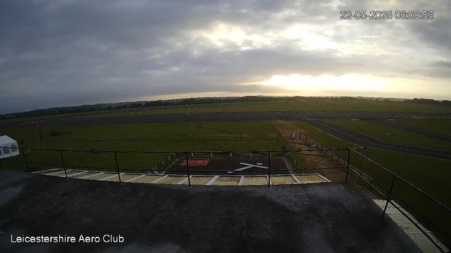 An outdoor view from a webcam shows a cloudy sky with the sun breaking through the clouds near the horizon. In the foreground, there is a balcony or observation deck with a black railing. Below the deck, there are marked parking spaces and a helipad with a white arrow pointing left and an "H" symbol. The background features an open grassy field with some fencing, a running track, and a few small structures at a distance. The timestamp indicates the date and time as May 22, 2025, at 6:00 AM, and the location is identified as Leicestershire Aero Club.