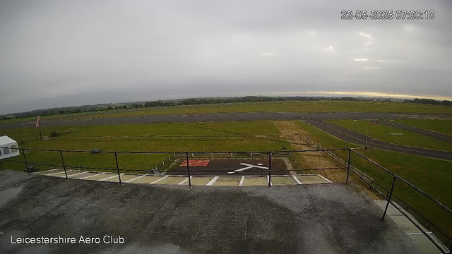 A view from a webcam at Leicesterhire Aero Club shows a runway and grassy area under a cloudy sky. The scene appears foggy or overcast, with a timestamp of 22-05-2025 at 07:00:10. The foreground includes a metal railing and a helipad marked with an "H" in white.