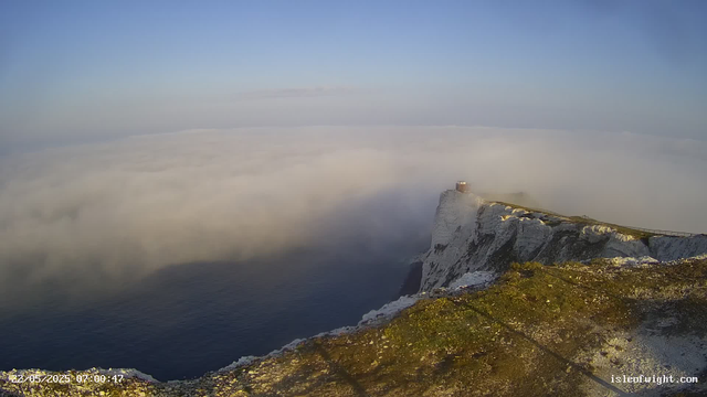 A view of white chalk cliffs on the coast, partially covered in moss and grass, with a small building or observatory at the edge of the cliff. Thick fog or low clouds hover over the ocean below, obscuring the water from view. The sky above is clear with a light blue hue, and the image appears to be taken at sunrise or early morning. The timestamp on the bottom left indicates the date and time as 22/05/2023 at 07:00:47.