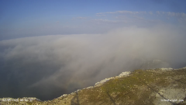 A view from a mountain webcam shows a rocky, moss-covered foreground with a thick layer of clouds below, partially obscuring the landscape. The sky above is mostly clear with some scattered clouds, and the scene is bathed in daylight. The timestamp in the lower left corner reads "22/05/2025 08:00:32," and the website "isleofwight.com" is visible in the lower right corner.
