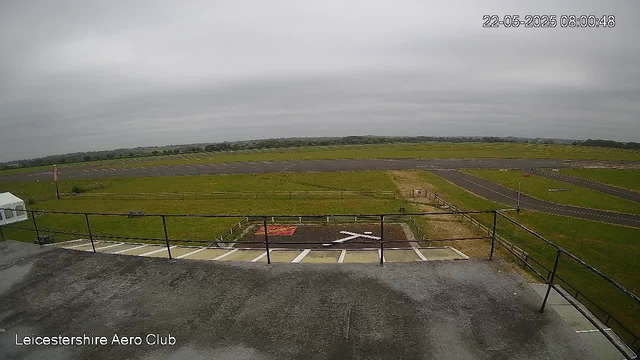 An empty parking area with white lines and an airplane parking space marked with an "X" at Leicestershire Aero Club. Beyond the parking lot, there is a grassy field with a runway and taxiways stretching into the distance under a cloudy sky. The photo has a timestamp showing "22-05-2025 08:00:48" in the upper right corner.