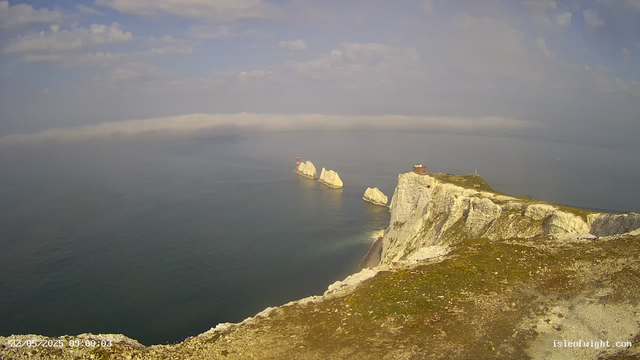 A scenic view of white chalk cliffs extending into the ocean, topped with grassy patches and a walkway, with large white rock formations jutting out of the sea in the distance. The sky is partly cloudy with blue patches, and the calm water reflects the sky above.