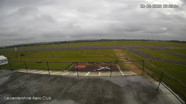 An outdoor view from a webcam showing an overcast sky with gray clouds. In the foreground, there is a concrete surface with a metal railing, and several marked parking spaces, including a red marked area. Beyond the railing, there is a grassy field with a small paved area and a runway or track extending into the distance. The image is labeled "Leicestershire Aero Club" in the bottom left corner, and the timestamp in the top right corner reads "22-05-2025 08:59:54."