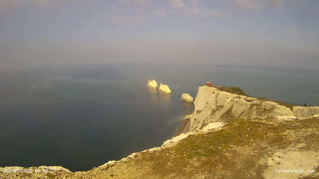 A view of white chalk cliffs by the seaside with a small lighthouse and a building at the top. Several large white rock formations are visible offshore in the calm blue-gray water. The sky is mostly clear with a few scattered clouds. The image appears to be taken during daytime.