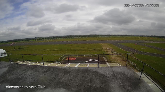 A wide view of an open outdoor space at an airport shows a grassy area with a small paved section in the foreground, marked with a white arrow and red paint. There is a metal fence running horizontally across the scene, with parking spaces visible at the bottom left corner. The sky is mostly cloudy with patches of blue, and the landscape extends into the distance with a runway and some low hills or structures on the horizon. The timestamp on the image indicates the date and time as May 22, 2025, at 10:00:23. The bottom left corner displays the text "Leicestershire Aero Club."