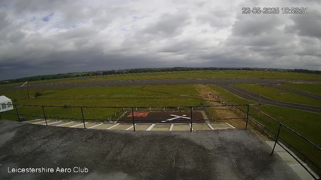 A view of an airfield with a grassy landscape extending into the distance under a cloudy sky. The foreground shows a concrete platform with a metal railing, and there are marked parking spaces and a small ramp leading down. The runway is visible to the right, with a clear path for aircraft takeoff and landing. The timestamp on the image reads "22-05-2025 10:59:51," and the bottom left corner has the text "Leicestershire Aero Club."
