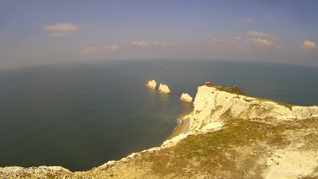 A scenic view of white chalk cliffs along a coastline, with a small building and a lighthouse at the top. The sea stretches out into the horizon with a few sailboats and ships visible. The sky is mostly clear with some scattered clouds.