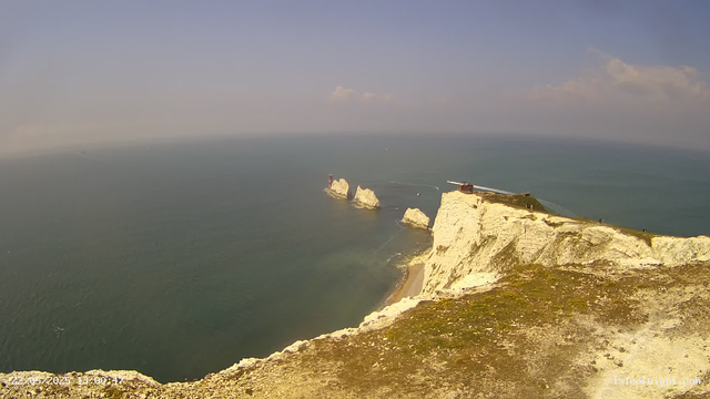 A view from a webcam shows white chalk cliffs extending into the ocean, with several large rock formations in the water nearby. At the top of the cliffs, there is a small building and a lighthouse, along with other structures. The sea is calm, and the sky is mostly clear with a few scattered clouds, creating a bright and sunny atmosphere.
