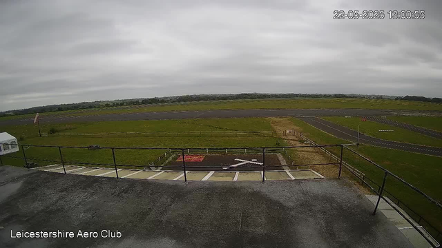 A wide view of an airfield with a runway in the distance, grassy areas on either side, and a cloudy sky overhead. The photo appears to be taken from a balcony or observation deck at Leicestershire Aero Club, with a railing visible in the foreground. There are marked parking spaces and a helipad in the lower part of the image. The timestamp shows the date as May 22, 2025, and the time as 13:00:55.